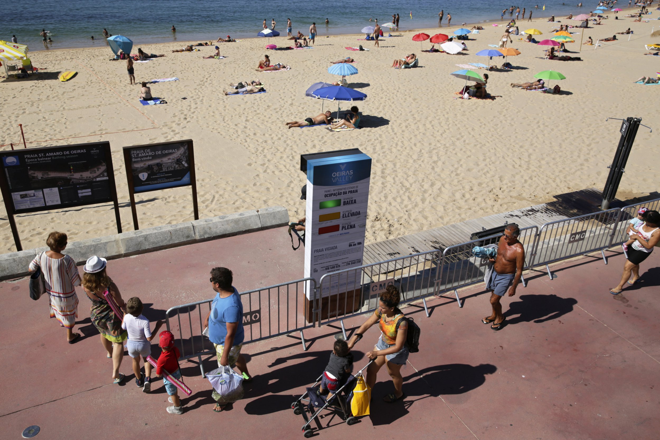 People arrive at the beach walk past a semaphore system that signals how crowded the beach is at any moment, in Oeiras, outside Lisbon, Wednesday, July 15, 2020. The system uses radar to count people entering the gates to the fenced beach to help beachgoers keep social distancing due to the coronavirus pandemic. (AP Photo/Armando Franca)